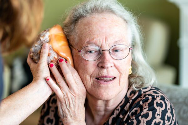oudere vrouw luistert tijdens het project kunstknuffel naar een grote schelp aan haar oor, vastgehouden door kunstdocent carmen